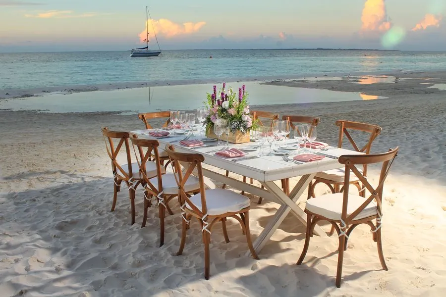 Mesa rectangular blanca en la playa con sillas crossback al atardecer