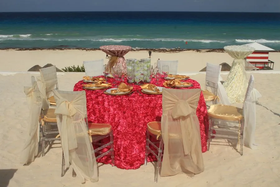 Mesa redonda en la playa con mantel rojo y fundas de silla